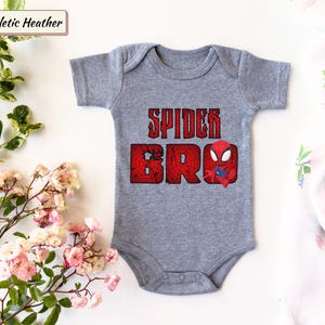 May include: A gray baby onesie with the text "SPIDER BRO" in distressed red lettering and a small Spider-Man graphic. The onesie is made of soft material and is designed for comfort.