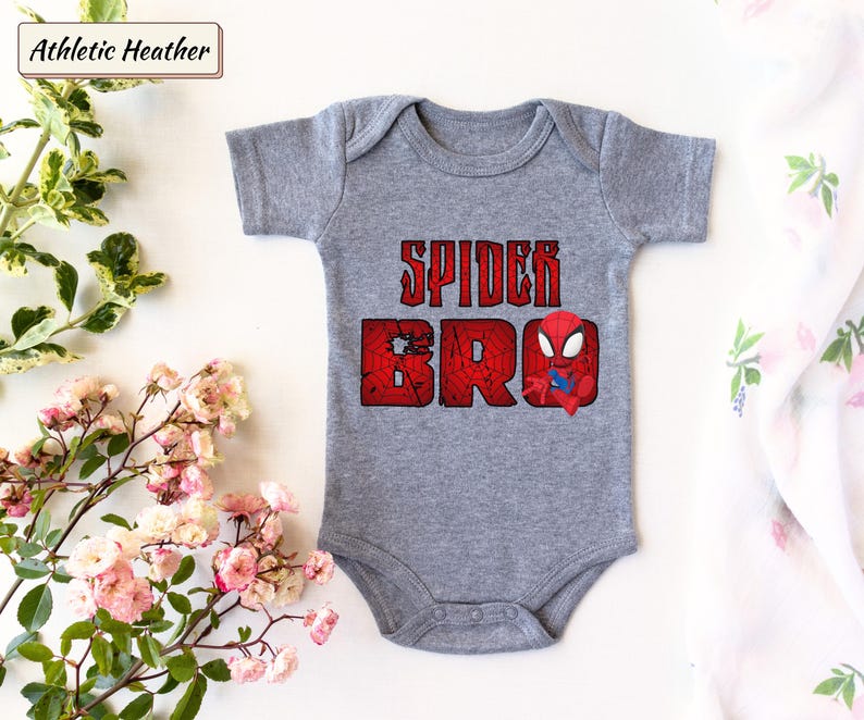 May include: A gray baby onesie with the text "SPIDER BRO" in distressed red lettering and a small Spider-Man graphic. The onesie is made of soft material and is designed for comfort.