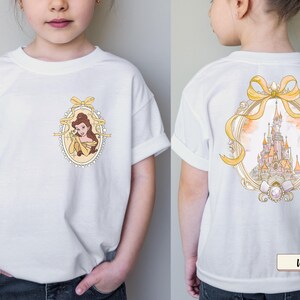 Op de afbeelding: Een wit T-shirt met korte mouwen met een kleine ovale afbeelding op de borst met een bruinharig personage in een gele jurk in een gouden lijst, en een grote afbeelding op de achterkant van een kasteel in een gouden lijst met een geel lint.