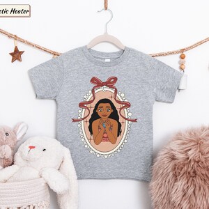 May include: An athletic heather gray short-sleeve t-shirt featuring a graphic print of a character with long dark hair inside an oval frame decorated with a red ribbon bow. The character is holding a blue stone pendant against a light background.