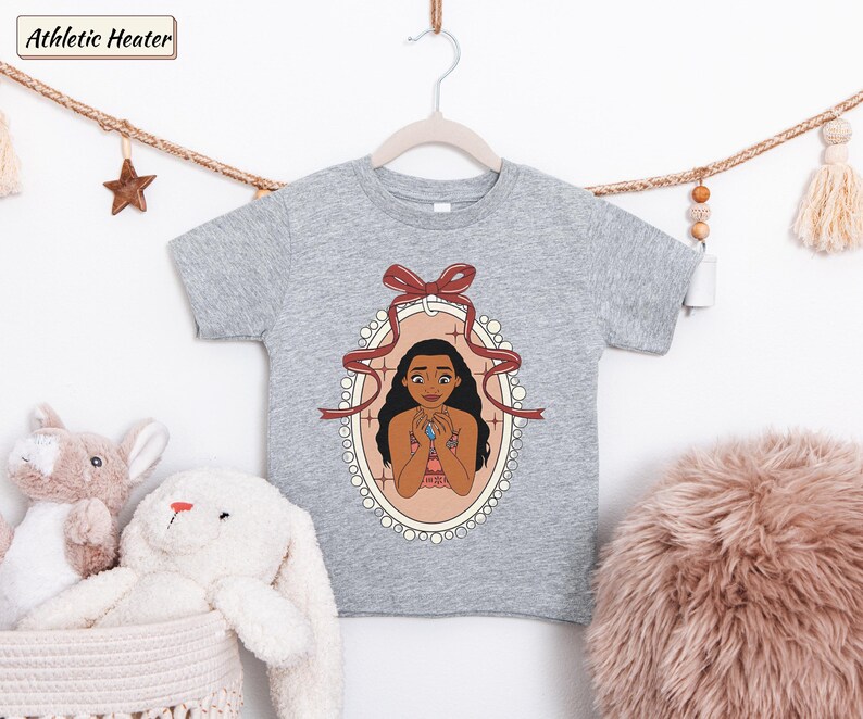 May include: An athletic heather gray short-sleeve t-shirt featuring a graphic print of a character with long dark hair inside an oval frame decorated with a red ribbon bow. The character is holding a blue stone pendant against a light background.