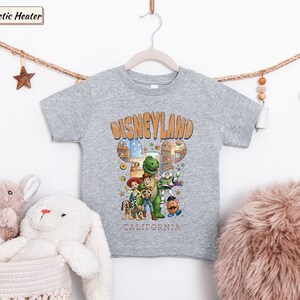 May include: A gray t-shirt featuring a Disneyland graphic with cartoon characters. The design includes the text "Disneyland California" in an orange, vintage-style font. The shirt is hanging on a wooden hanger.