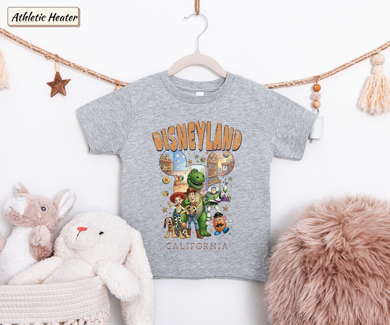 May include: A gray t-shirt featuring a Disneyland graphic with cartoon characters. The design includes the text "Disneyland California" in an orange, vintage-style font. The shirt is hanging on a wooden hanger.