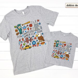 May include: Two athletic heather gray t-shirts featuring a colorful graphic print of American icons including the Statue of Liberty, a bald eagle, the Capitol building, a yellow taxi, and the text 1776 HAPPY 250 AMERICA 2026 with stars.