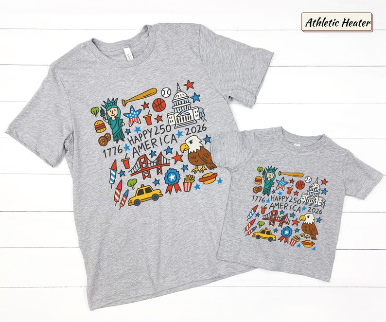 May include: Two athletic heather gray t-shirts featuring a colorful graphic print of American icons including the Statue of Liberty, a bald eagle, the Capitol building, a yellow taxi, and the text 1776 HAPPY 250 AMERICA 2026 with stars.