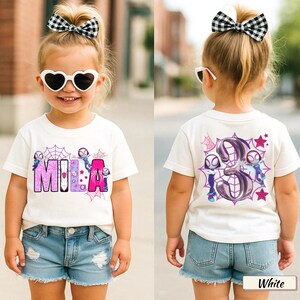 K&ouml;nnte beinhalten: Wei&szlig;es T-Shirt mit dem Namen "MILA" in rosa und lila Buchstaben, mit Cartoon-Figuren und Spinnennetzen. Auf der R&uuml;ckseite des Shirts ist die Zahl "3" mit Cartoon-Figuren, Spinnennetzen und Sternen abgebildet. Das Kind tr&auml;gt Jeansshorts und herzf&ouml;rmige Sonnenbrillen.