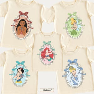 May include: Five cream-colored short-sleeve t-shirts featuring oval frames with scalloped edges. Each frame contains a different cartoon character: Moana with a red bow, Tinker Bell with a green bow, Ariel with a teal bow, Snow White with a blue bow, and Cinderella with a light blue bow.