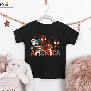 May include: A black short-sleeve t-shirt featuring a graphic of three cartoon spider-themed characters in front of an American flag with fireworks. The word AMERICA is printed below in a bold, dotted font with red, white, and blue colors.