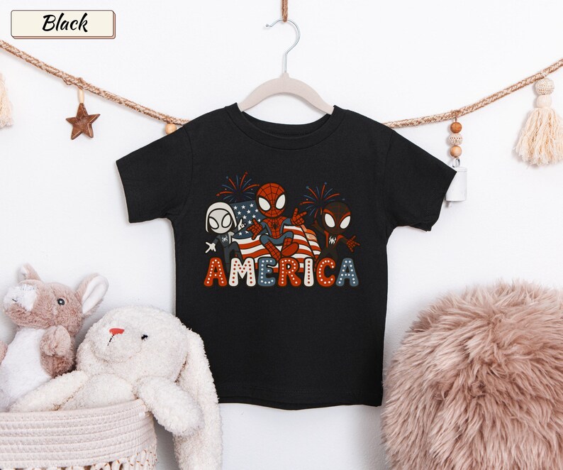 May include: A black short-sleeve t-shirt featuring a graphic of three cartoon spider-themed characters in front of an American flag with fireworks. The word AMERICA is printed below in a bold, dotted font with red, white, and blue colors.