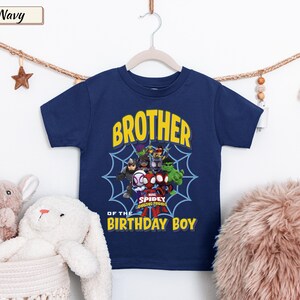 May include: A navy blue short-sleeve t-shirt featuring a graphic of Marvel Spidey and His Amazing Friends characters centered over a blue spiderweb. The text reads BROTHER OF THE BIRTHDAY BOY in yellow block letters above and below the characters.