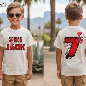 May include: Off-white t-shirt with the text "SPIDER JACK" in red and a Spider-Man graphic. The back of the shirt features a large red number 7 with a spider web and a Spider-Man figure. The shirt is worn by a child.