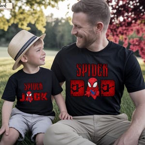 May include: Black t-shirts with red Spider-Man graphics. One shirt says "Spider Dad" with a Spider-Man character, and the other says "Spider Jack" with a Spider-Man character. A child wears a straw hat and gray shorts.