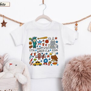 May include: A white short-sleeve t-shirt featuring a colorful graphic print of American icons including the Statue of Liberty, a bald eagle, a taxi, and the text "HAPPY 250 AMERICA 1776 2026" surrounded by stars, sports equipment, and food items.
