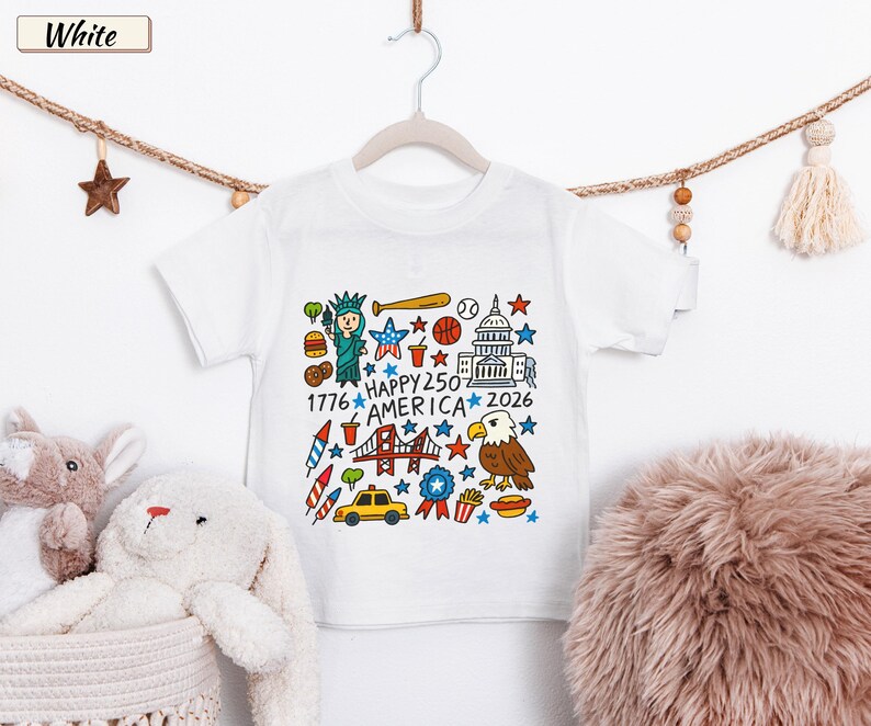 May include: A white short-sleeve t-shirt featuring a colorful graphic print of American icons including the Statue of Liberty, a bald eagle, a taxi, and the text "HAPPY 250 AMERICA 1776 2026" surrounded by stars, sports equipment, and food items.