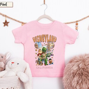 May include: Pink short-sleeved t-shirt featuring a Disneyland graphic with cartoon characters. The design includes the word "Disneyland" in a brown font, with "California" below. The shirt is hanging on a white hanger.