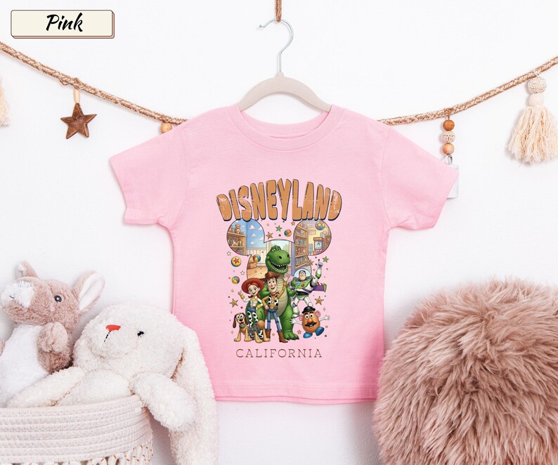 May include: Pink short-sleeved t-shirt featuring a Disneyland graphic with cartoon characters. The design includes the word "Disneyland" in a brown font, with "California" below. The shirt is hanging on a white hanger.