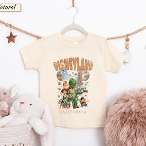 May include: Cream-colored t-shirt featuring the word "Disneyland" in a brown, retro font. The shirt has a graphic of Disney characters. Below the graphic, the word "California" is printed. The shirt is hanging on a white hanger.