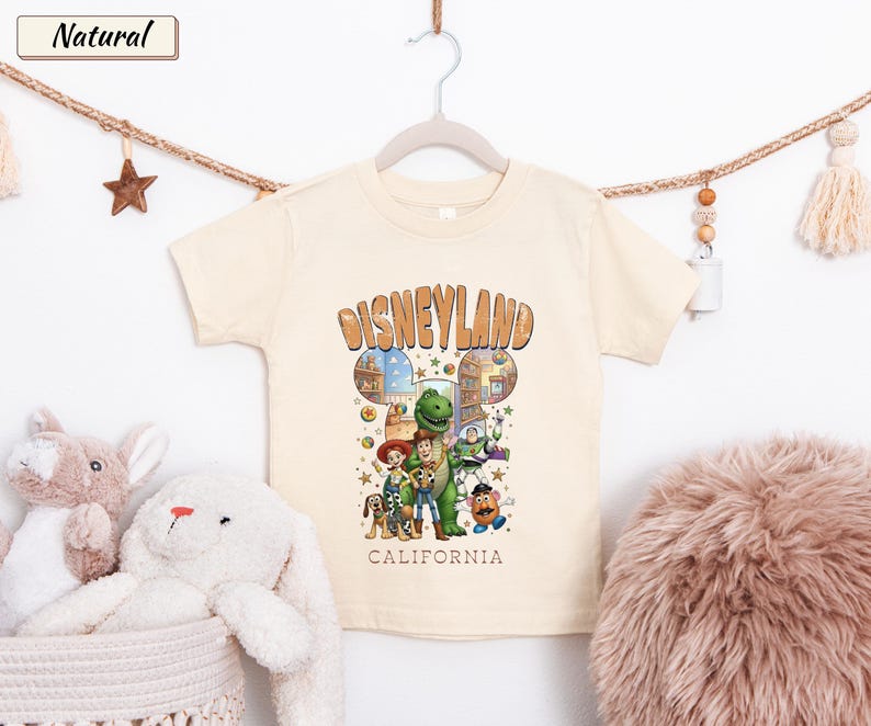 May include: Cream-colored t-shirt featuring the word "Disneyland" in a brown, retro font. The shirt has a graphic of Disney characters. Below the graphic, the word "California" is printed. The shirt is hanging on a white hanger.