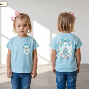 Op de afbeelding: Een ijsblauw T-shirt met korte mouwen, voorzien van een kleine ovale afbeelding van een blond personage op de linkerborst en een grote, gedetailleerde illustratie van een kasteel omringd door een blauw lint en een gouden rand op de achterkant.
