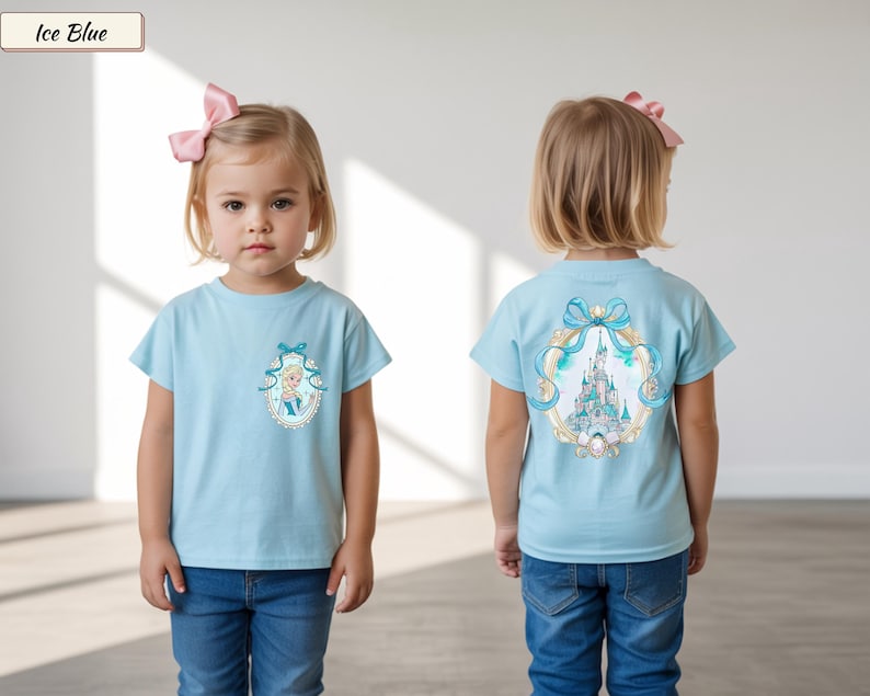 Op de afbeelding: Een ijsblauw T-shirt met korte mouwen, voorzien van een kleine ovale afbeelding van een blond personage op de linkerborst en een grote, gedetailleerde illustratie van een kasteel omringd door een blauw lint en een gouden rand op de achterkant.