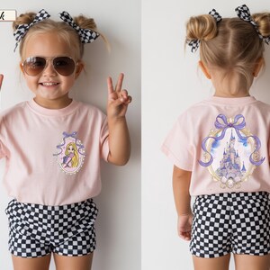 Op de afbeelding: Een lichtroze t-shirt met een kleine afbeelding van een blond personage in een paarse jurk op de linkerborst en een grote afbeelding van een kasteel omlijst door een paarse strik op de achterkant, gecombineerd met zwart-wit geblokte shorts.