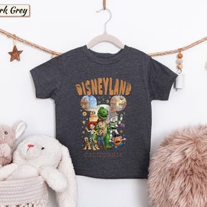 May include: Dark grey t-shirt featuring a Disneyland design with cartoon characters. The word "Disneyland" is in orange, with the word "California" below. The shirt is hanging on a wooden hanger.