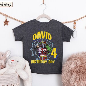 May include: A dark grey short-sleeve t-shirt featuring a graphic of Marvel Spidey and His Amazing Friends characters inside a spiderweb. The text reads DAVID, 4, and BIRTHDAY BOY in yellow and blue lettering on the front of the garment.