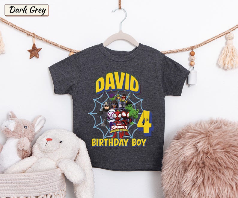 May include: A dark grey short-sleeve t-shirt featuring a graphic of Marvel Spidey and His Amazing Friends characters inside a spiderweb. The text reads DAVID, 4, and BIRTHDAY BOY in yellow and blue lettering on the front of the garment.