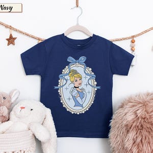 May include: A navy blue short-sleeved t-shirt featuring a graphic print of Cinderella inside an oval, pearl-bordered frame. The design includes a light blue ribbon bow at the top of the frame and small star accents surrounding the character illustration.