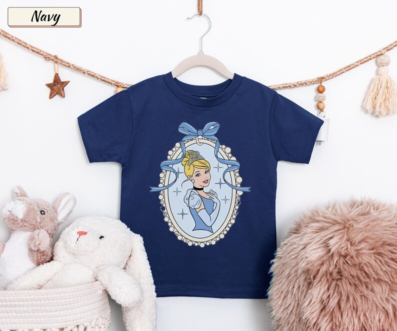 May include: A navy blue short-sleeved t-shirt featuring a graphic print of Cinderella inside an oval, pearl-bordered frame. The design includes a light blue ribbon bow at the top of the frame and small star accents surrounding the character illustration.