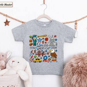May include: A heather grey t-shirt featuring a colorful graphic print with the text "HAPPY 250 AMERICA 1776 2026". The design includes illustrations of the Statue of Liberty, a bald eagle, the Golden Gate Bridge, a taxi, food items, and stars.