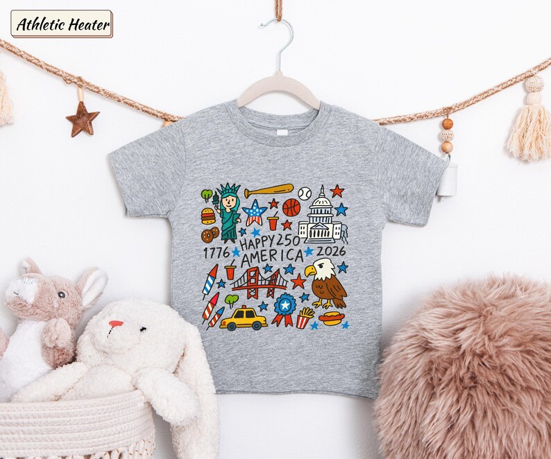 May include: A heather grey t-shirt featuring a colorful graphic print with the text "HAPPY 250 AMERICA 1776 2026". The design includes illustrations of the Statue of Liberty, a bald eagle, the Golden Gate Bridge, a taxi, food items, and stars.