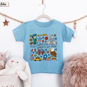 May include: An ice blue short-sleeve t-shirt featuring a colorful graphic print of American icons including the Statue of Liberty, a bald eagle, a baseball, a taxi, and the text "HAPPY 250 AMERICA 1776 2026" arranged in a square pattern.