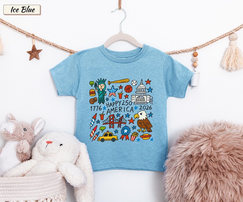 May include: An ice blue short-sleeve t-shirt featuring a colorful graphic print of American icons including the Statue of Liberty, a bald eagle, a baseball, a taxi, and the text "HAPPY 250 AMERICA 1776 2026" arranged in a square pattern.