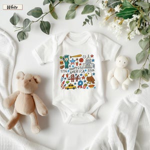 May include: A white short-sleeved baby bodysuit featuring a colorful graphic print of American icons including the Statue of Liberty, a bald eagle, the Capitol building, and a yellow taxi, with the text "HAPPY 250 AMERICA 1776 2026".