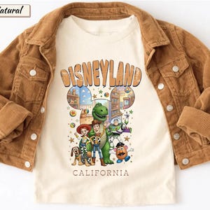 May include: A tan corduroy jacket over a cream-colored t-shirt featuring a Disneyland graphic with cartoon characters. The shirt has the words "Disneyland California" in orange. The jacket has button closures and pockets.