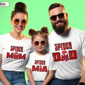 May include: Three people wearing white t-shirts with red and black Spider-Man themed designs. The shirts read "Spider Mom", "Spider Mia", and "Spider Dad". The background is a solid green color.