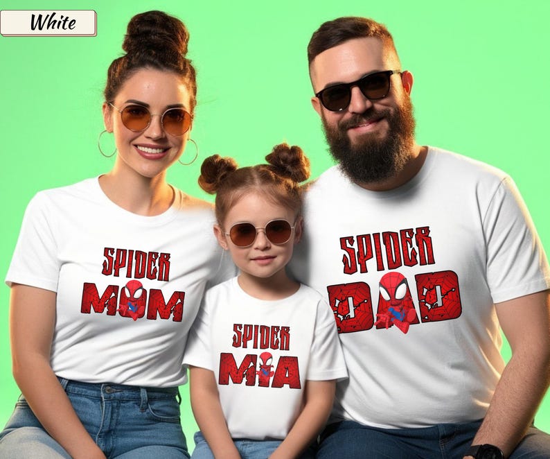 May include: Three people wearing white t-shirts with red and black Spider-Man themed designs. The shirts read "Spider Mom", "Spider Mia", and "Spider Dad". The background is a solid green color.