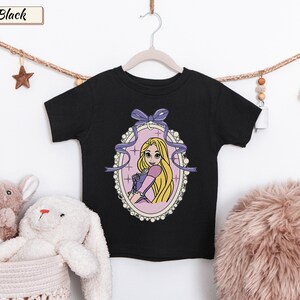 May include: A black short-sleeve t-shirt featuring a graphic print of a blonde character with long hair in a purple dress, framed by an oval border with a purple bow at the top. The text "Black" appears in a small rectangular box at the top left.