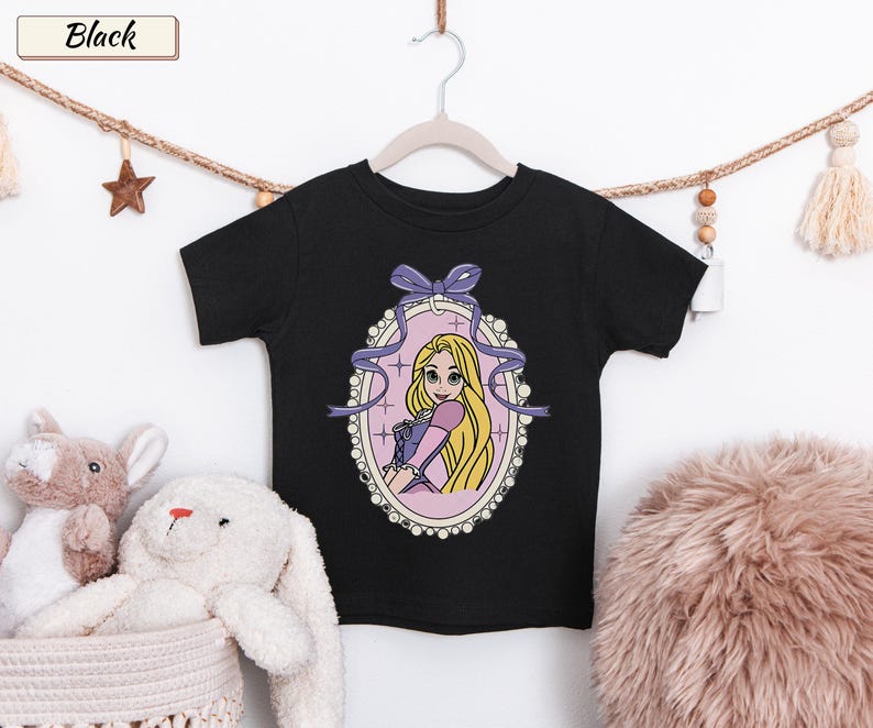 May include: A black short-sleeve t-shirt featuring a graphic print of a blonde character with long hair in a purple dress, framed by an oval border with a purple bow at the top. The text "Black" appears in a small rectangular box at the top left.