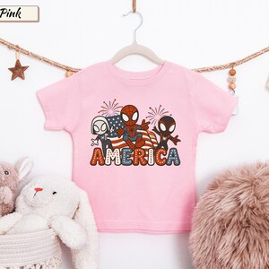 May include: A light pink short-sleeve t-shirt featuring a graphic print of three cartoon spider-themed characters in front of an American flag and fireworks. The word AMERICA is printed in large, bold, red, white, and blue dotted letters below.