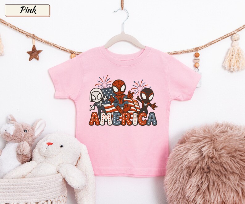 May include: A light pink short-sleeve t-shirt featuring a graphic print of three cartoon spider-themed characters in front of an American flag and fireworks. The word AMERICA is printed in large, bold, red, white, and blue dotted letters below.