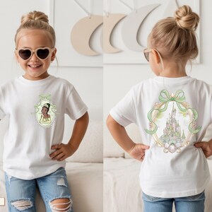 Op de afbeelding: Een wit t-shirt met korte mouwen en een kleine afbeelding op de linkerborst van een prinses in een groene jurk binnen een ovaal kader. De achterkant van het shirt toont een grote illustratie van een kasteel in een sierlijk ovaal kader met een groene strik.