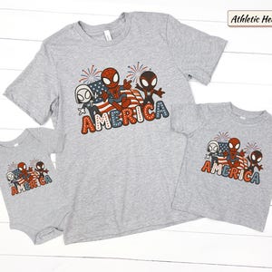 May include: An athletic heather gray t-shirt and matching baby bodysuit featuring a graphic of three cartoon spider characters, a waving American flag, and fireworks. The word AMERICA is printed in large, dotted, red, white, and blue letters.