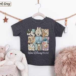 May include: A dark grey short-sleeve t-shirt featuring a grid of nine squares containing illustrations of various Disney characters and a castle. Below the grid, the text Walt Disney World is printed in a white script font on the front of the shirt.