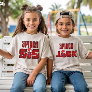May include: Two children wearing beige t-shirts with red and black Spider-Man graphics. The shirts have the words "Spider" and "SS" or "Jack" printed on them. They are sitting on a white bench, smiling at the camera.