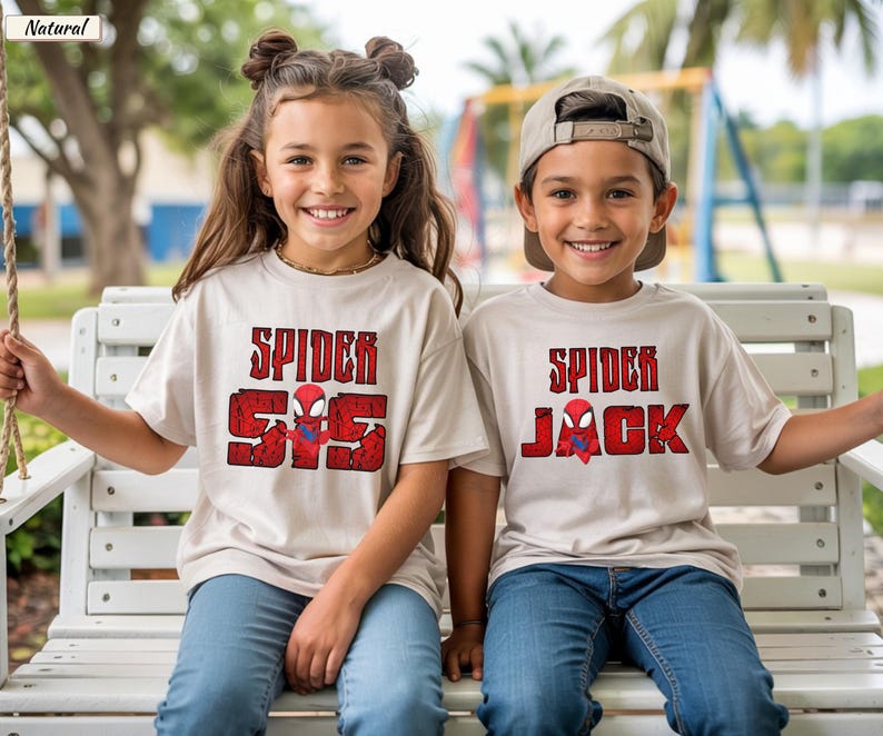 May include: Two children wearing beige t-shirts with red and black Spider-Man graphics. The shirts have the words "Spider" and "SS" or "Jack" printed on them. They are sitting on a white bench, smiling at the camera.