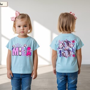 K&ouml;nnte beinhalten: Hellblaues T-Shirt mit dem Namen "MIA" in rosa und lila Buchstaben, mit einem Spinnennetz und Zeichentrickfiguren. Auf der R&uuml;ckseite des Shirts ist eine gro&szlig;e Zahl "3" mit Zeichentrickfiguren und Sternen abgebildet. Das Shirt wird von einem kleinen Kind getragen.