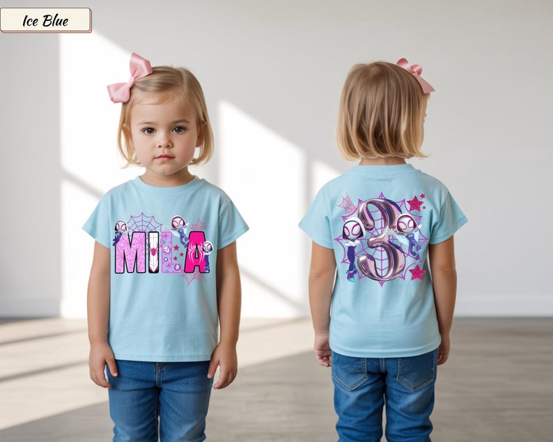 K&ouml;nnte beinhalten: Hellblaues T-Shirt mit dem Namen "MIA" in rosa und lila Buchstaben, mit einem Spinnennetz und Zeichentrickfiguren. Auf der R&uuml;ckseite des Shirts ist eine gro&szlig;e Zahl "3" mit Zeichentrickfiguren und Sternen abgebildet. Das Shirt wird von einem kleinen Kind getragen.