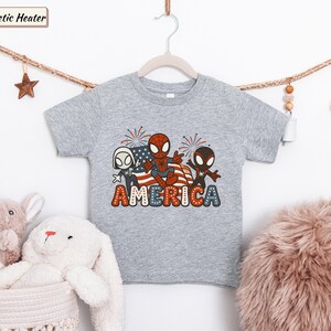 May include: A heather gray short-sleeve t-shirt featuring a graphic of three Spider-Man characters in front of an American flag with fireworks. The word AMERICA is printed below in large, bold, red, white, and blue polka-dotted lettering.